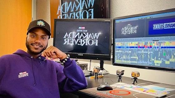 Alain sits at a desk with multiple monitors that read “Wakanda Forever” and “Marvel Studios.他穿着一件紫色卫衣.